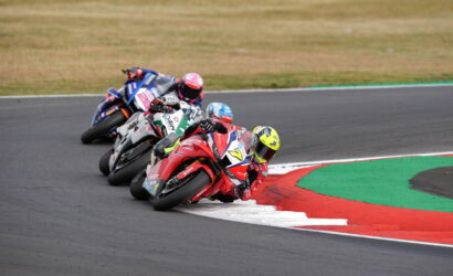 MotoGP-style race: four riders lean into a tight right-hand curve on a paved track with red, white, and green run-off areas ahead.