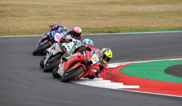 MotoGP-style race: four riders lean into a tight right-hand curve on a paved track with red, white, and green run-off areas ahead.