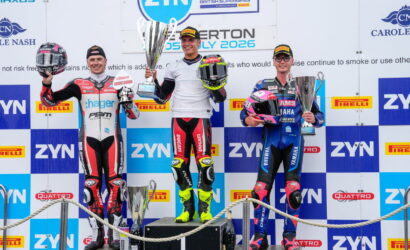 Three motorcycle racers stand on a podium holding trophies, celebrating a race finish with sponsor banners in the background.