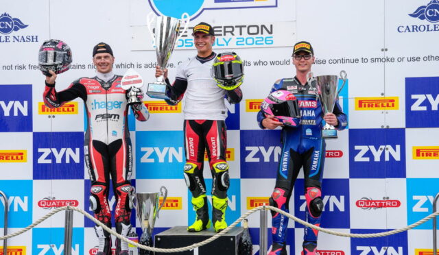 Three motorcycle racers stand on a podium holding trophies, celebrating a race finish with sponsor banners in the background.
