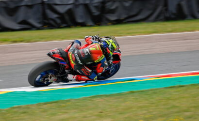 Motorcycle racer in red suit leaning into a sharp turn on a racing track, colorful helmet and bike decals visible.