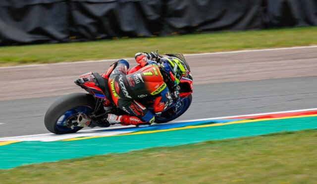 Motorcycle racer in red suit leaning into a sharp turn on a racing track, colorful helmet and bike decals visible.