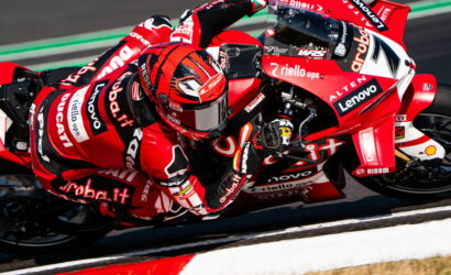 MotoGP rider in a red racing suit leaning into a turn on a red Ducati race bike with sponsor logos visible on the bike and suit.