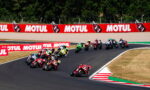 Motorcycle race on a curving track with a pack of riders leaning into a turn and red Motul banners along the outside wall.