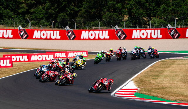 Motorcycle race on a curving track with a pack of riders leaning into a turn and red Motul banners along the outside wall.