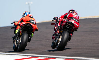 Two motorcycle racers in red gear lean into a curb on a race track, side by side during a high-speed duel.
