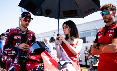 Two motorcycle racers in red racing suits stand at a pit area while a woman in a red outfit holds a large umbrella over them, adjusting a helmet on the bike.