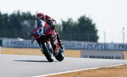 Professional motorcycle racer in red leathers leaning into a turn on a red race bike with sponsor logos on a sunny track.