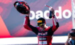 Race driver in red-and-black suit lifts helmet in celebration on a podium, with sponsor logos visible in the background.