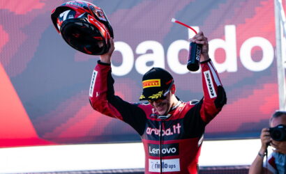 Motorsport racer in red and black suit raises helmet and a champagne bottle in celebration on a podium.