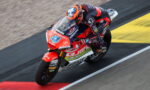 Motocycle racer in red-and-black suit leaning into a turn on a red racing bike on track surface.