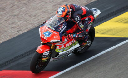 Motocycle racer in red-and-black suit leaning into a turn on a red racing bike on track surface.