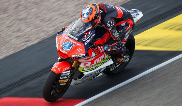 Motocycle racer in red-and-black suit leaning into a turn on a red racing bike on track surface.