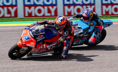 Two motorcycle racers lean into a left turn on a sunlit track, red and blue bikes close together with Liqui Moly banners in the background.
