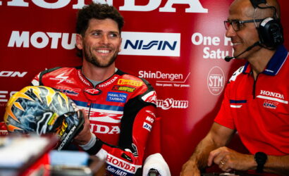 Professional motorcycle racer in a red Honda racing suit holds his helmet and smiles, as a team member with a headset chats beside him in the pit area.