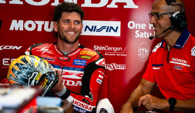 Professional motorcycle racer in a red Honda racing suit holds his helmet and smiles, as a team member with a headset chats beside him in the pit area.