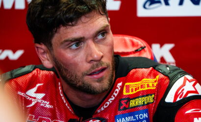 Close-up of a male MotoGP rider in a red racing suit, seated at a press event, looking off to the side.