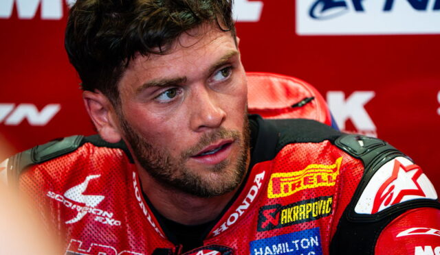Close-up of a male MotoGP rider in a red racing suit, seated at a press event, looking off to the side.