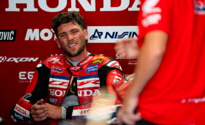 MotoGP rider in a red Honda racing suit sits in a red pit-area backdrop with sponsor logos, looking tired or frustrated during a break in the session.