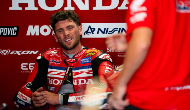 MotoGP rider in a red Honda racing suit sits in a red pit-area backdrop with sponsor logos, looking tired or frustrated during a break in the session.