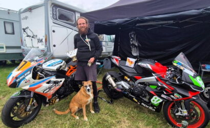 Bearded man in a black hoodie stands between two race motorcycles with a brown dog at his feet under a canopy on a grassy area.