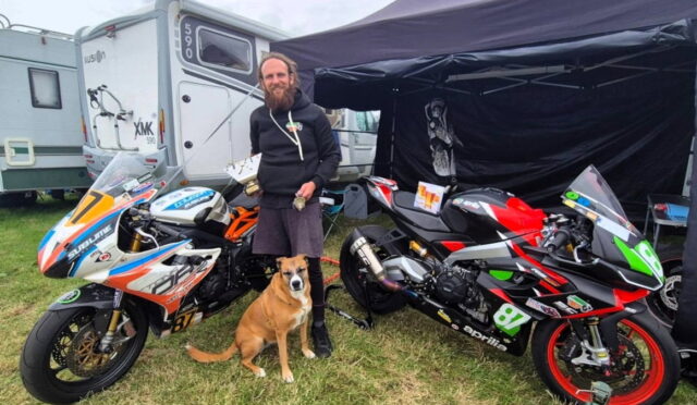 Bearded man in a black hoodie stands between two race motorcycles with a brown dog at his feet under a canopy on a grassy area.