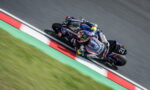 Motocycle racer in blue Yamaha suit leans into a sharp turn on a race track, motorcycles trailing closely behind on a curbed red-white-green section, motion blur in the background.