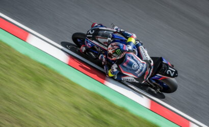 Motocycle racer in blue Yamaha suit leans into a sharp turn on a race track, motorcycles trailing closely behind on a curbed red-white-green section, motion blur in the background.