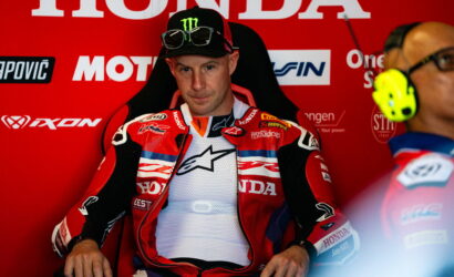 Motorcycle racer in red Honda racing suit seated in a red pit area, with sponsor logos in the background and a teammate wearing yellow ear protection nearby