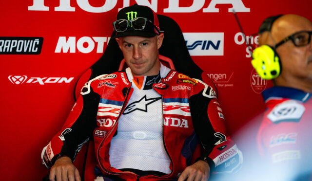 Motorcycle racer in red Honda racing suit seated in a red pit area, with sponsor logos in the background and a teammate wearing yellow ear protection nearby