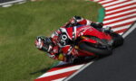 Motorcycle racer in red and white gear leans into a corner on a racetrack, bike number 30.