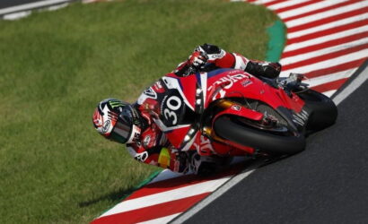 Motorsport rider in red leathers leaning into a corner on a red racing motorcycle on a track edge with red-and-white curbing.