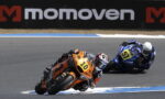 Two motorcycle racers lean into a turn on a race track; orange bike #10 ahead of blue bike #51 in pursuit.