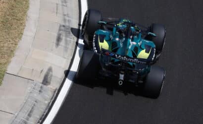 Top-down view of a teal Formula 1 car hugging the inside of a curving race track, with a concrete curb on the left and dark asphalt on the right.