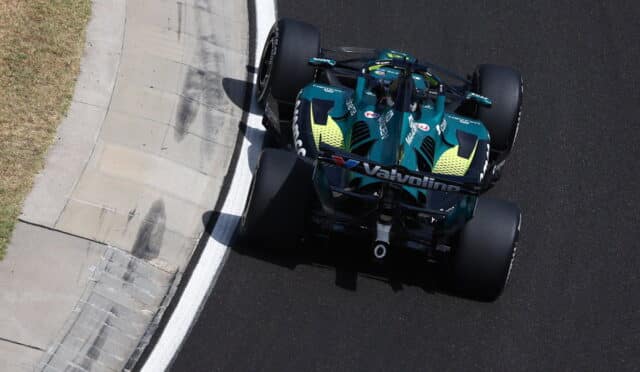 Top-down view of a teal Formula 1 car hugging the inside of a curving race track, with a concrete curb on the left and dark asphalt on the right.