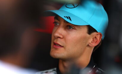 Close-up of a man wearing a bright blue cap with a Mercedes logo, looking off to the left in a thoughtful gaze.