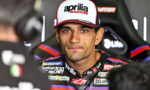 Motorcycle racer wearing an Aprilia Racing cap and colorful sponsor-studded suit, seated and looking at the camera during an interview setting