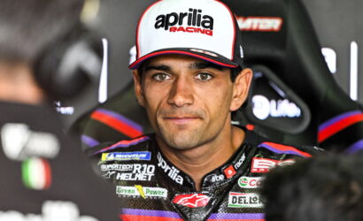 Motorcycle racer wearing an Aprilia Racing cap and colorful sponsor-studded suit, seated and looking at the camera during an interview setting