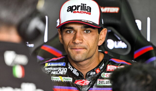 Motorcycle racer wearing an Aprilia Racing cap and colorful sponsor-studded suit, seated and looking at the camera during an interview setting
