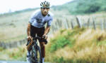 Male cyclist in a white jersey and helmet rides a road bike on a countryside road with a fence in the background, mid-race.