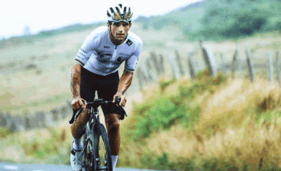 Male cyclist in a white jersey and helmet rides a road bike on a countryside road with a fence in the background, mid-race.