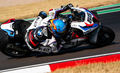 Two riders on a white, blue, and red race bike lean into a left-hand turn on a racetrack with red-and-white curbing visible in the foreground, speed evident.