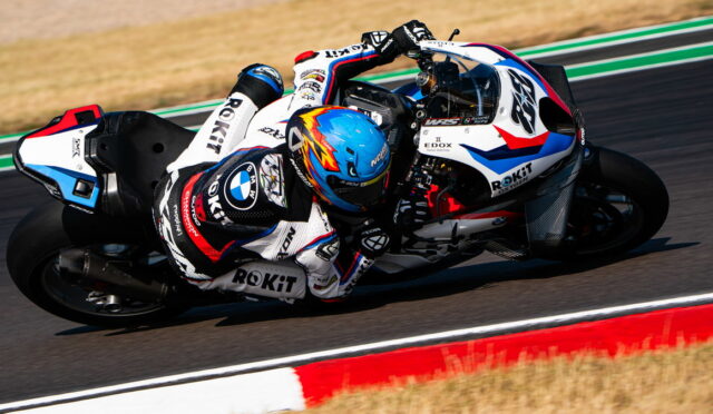 Two riders on a white, blue, and red race bike lean into a left-hand turn on a racetrack with red-and-white curbing visible in the foreground, speed evident.
