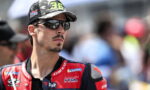 Racer wearing a red-and-blue Dainese racing suit and camouflage cap, sunglasses on, looking off to the side at a race event