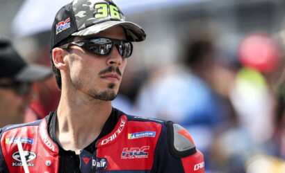 Racer wearing a red-and-blue Dainese racing suit and camouflage cap, sunglasses on, looking off to the side at a race event