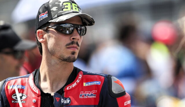 Racer wearing a red-and-blue Dainese racing suit and camouflage cap, sunglasses on, looking off to the side at a race event