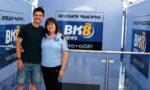Man and woman pose arm-in-arm in front of a blue Gresini Racing backdrop with BK8 News MotoGP logos; both smile at the camera.