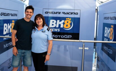 Man and woman pose arm-in-arm in front of a blue Gresini Racing backdrop with BK8 News MotoGP logos; both smile at the camera.