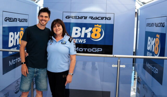 Man and woman pose arm-in-arm in front of a blue Gresini Racing backdrop with BK8 News MotoGP logos; both smile at the camera.