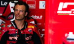 Male motorcyclist in a red Ducati racing suit sits in a sponsor-filled pit area, looking off to the side.
